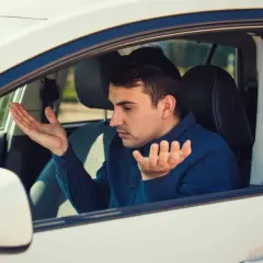 Homme frustré dans sa voiture qui tremble au point mort, mains levées en signe d'incompréhension.