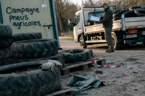 Un homme près d'un camion chargé de pneus agricoles. Un panneau indique "Campagne Pneus agricoles", un lieu où jeter des pneus usagés.
