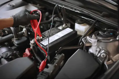 Main d'une personne portant un gant noir connectant des câbles de démarrage à la batterie d'une voiture qui s'éteint en roulant.