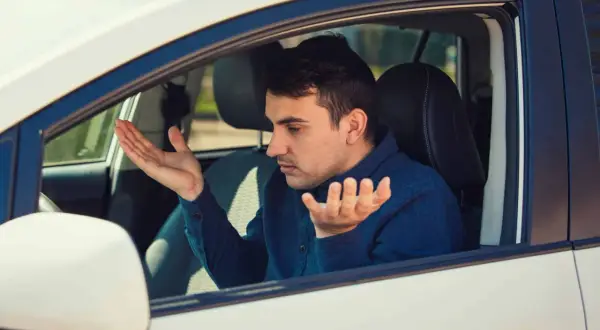 Homme frustré dans sa voiture qui tremble au point mort, mains levées en signe d'incompréhension.