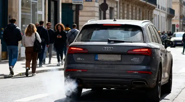 Une Audi émet une épaisse fumée blanche, signe d'une mauvaise odeur d'échappement diesel. Des passants marchent sur le trottoir.