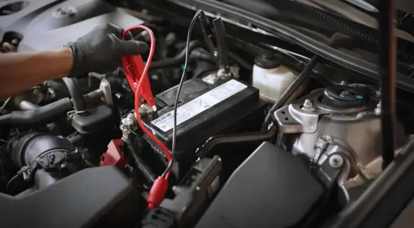 Main d'une personne portant un gant noir connectant des câbles de démarrage à la batterie d'une voiture qui s'éteint en roulant.