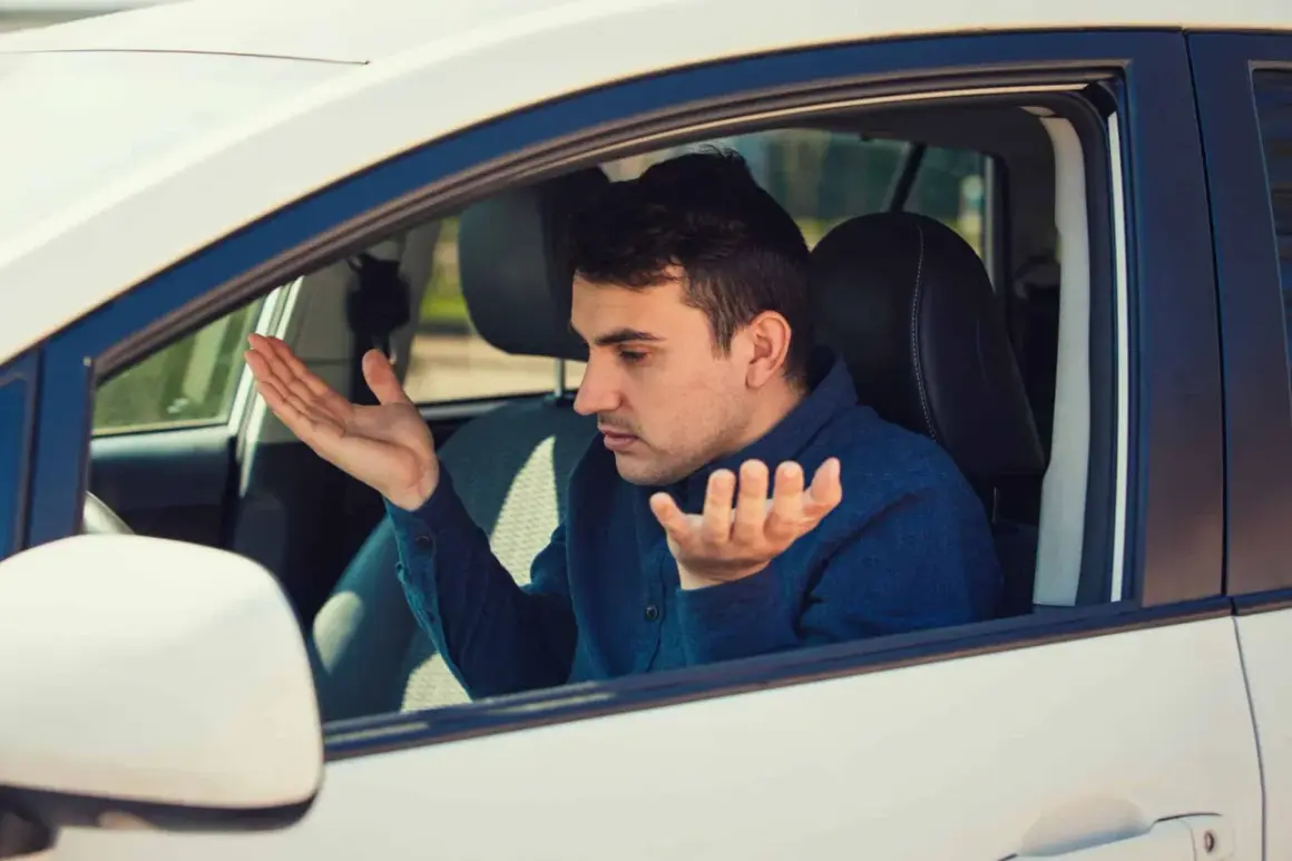 Homme frustré dans sa voiture qui tremble au point mort, mains levées en signe d'incompréhension.