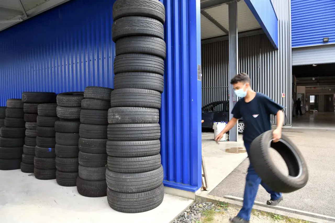 Un jeune homme porte un pneu usag&eacute;, pr&ecirc;t &agrave; le jeter. Des piles de pneus s'alignent le long d'un mur bleu.