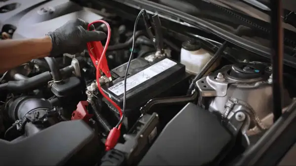 Main d'une personne portant un gant noir connectant des câbles de démarrage à la batterie d'une voiture qui s'éteint en roulant.
